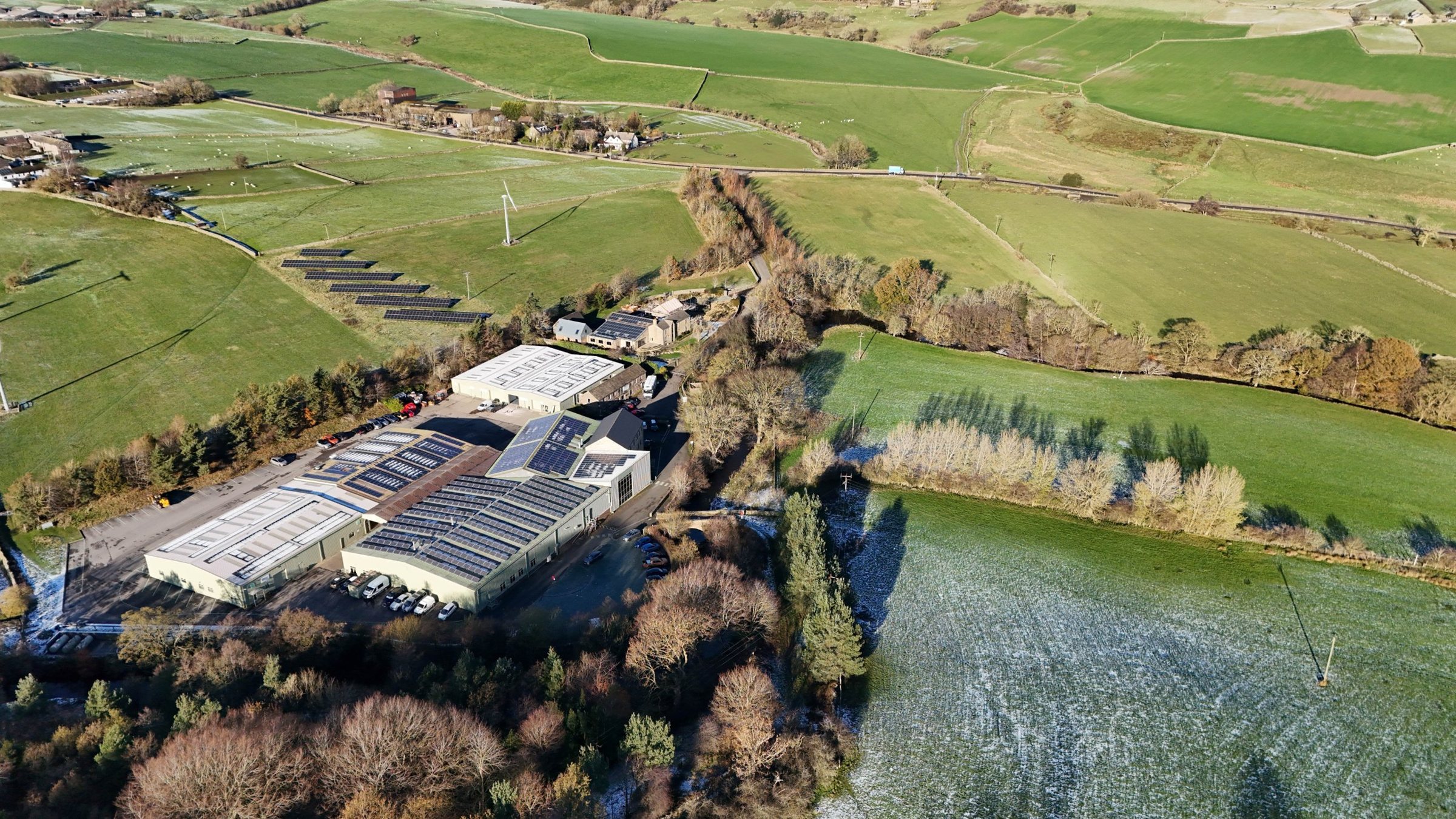 Aerial view of Bullhouse Mill in its valley, with solar panels on the roofs, a ground-mounted solar array and a wind turbine in the fields beyond