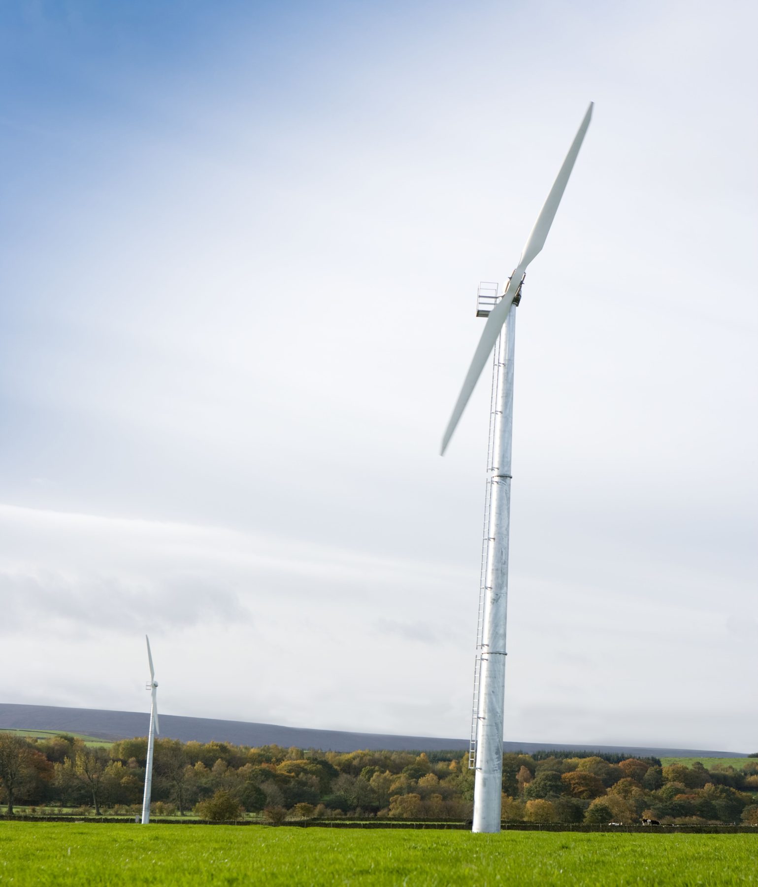 Two wind turbines in green fields, with autumn woodland and moorland behind
